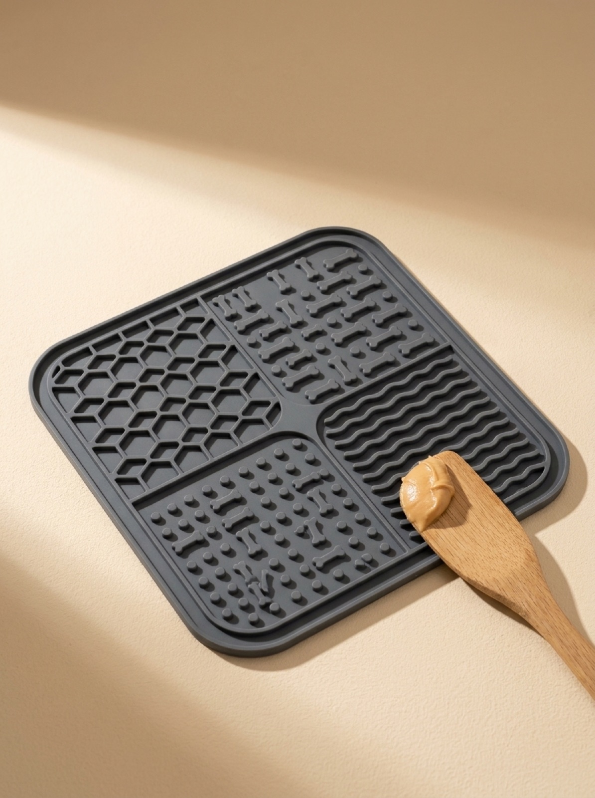 Tapis de léchage Naïa Sérénité en silicone gris anthracite et spatule en bois garnie de beurre de cacahuète, packshot sur fond crème